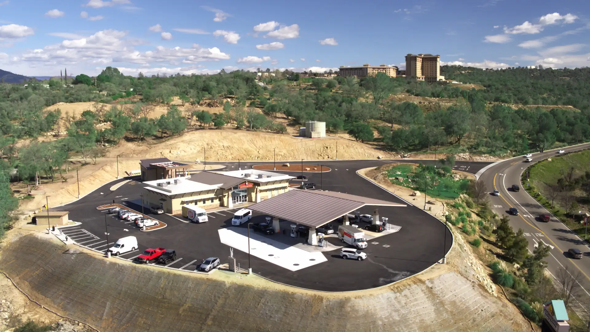 Aerial view of a gas station situated on a hill with a large, paved parking area. The gas station features a building with a light-colored roof and a canopy sheltering several fuel pumps. Multiple vehicles, including trucks and cars, are in the parking lot. Behind the gas station, there is a vast area of green vegetation and trees stretching into the distance. A winding road curves around the hill, with more vehicles visible. In the background, a large multi-story building is perched atop another hill. The sky is mostly clear with some scattered clouds.