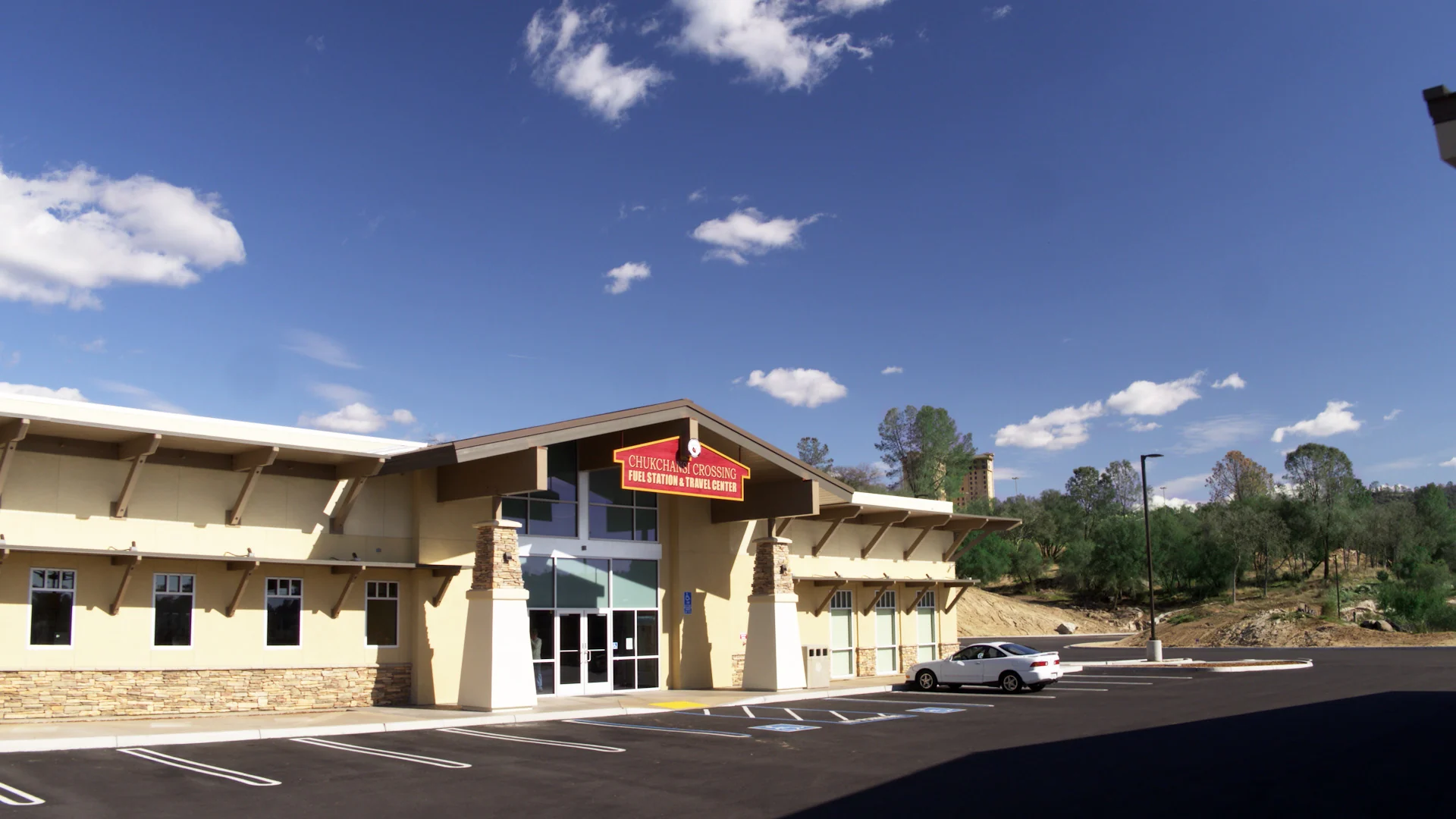 The image shows a modern building under a clear blue sky with a few scattered clouds. The structure is a single-story facility with cream-colored walls, stone accents, and large windows. A red and yellow sign on the building